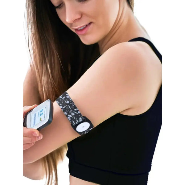 A woman using a smartphone app while wearing a Freestyle Libre 2 Armband with a patterned strap on her upper arm. The armband securely holds the sensor.